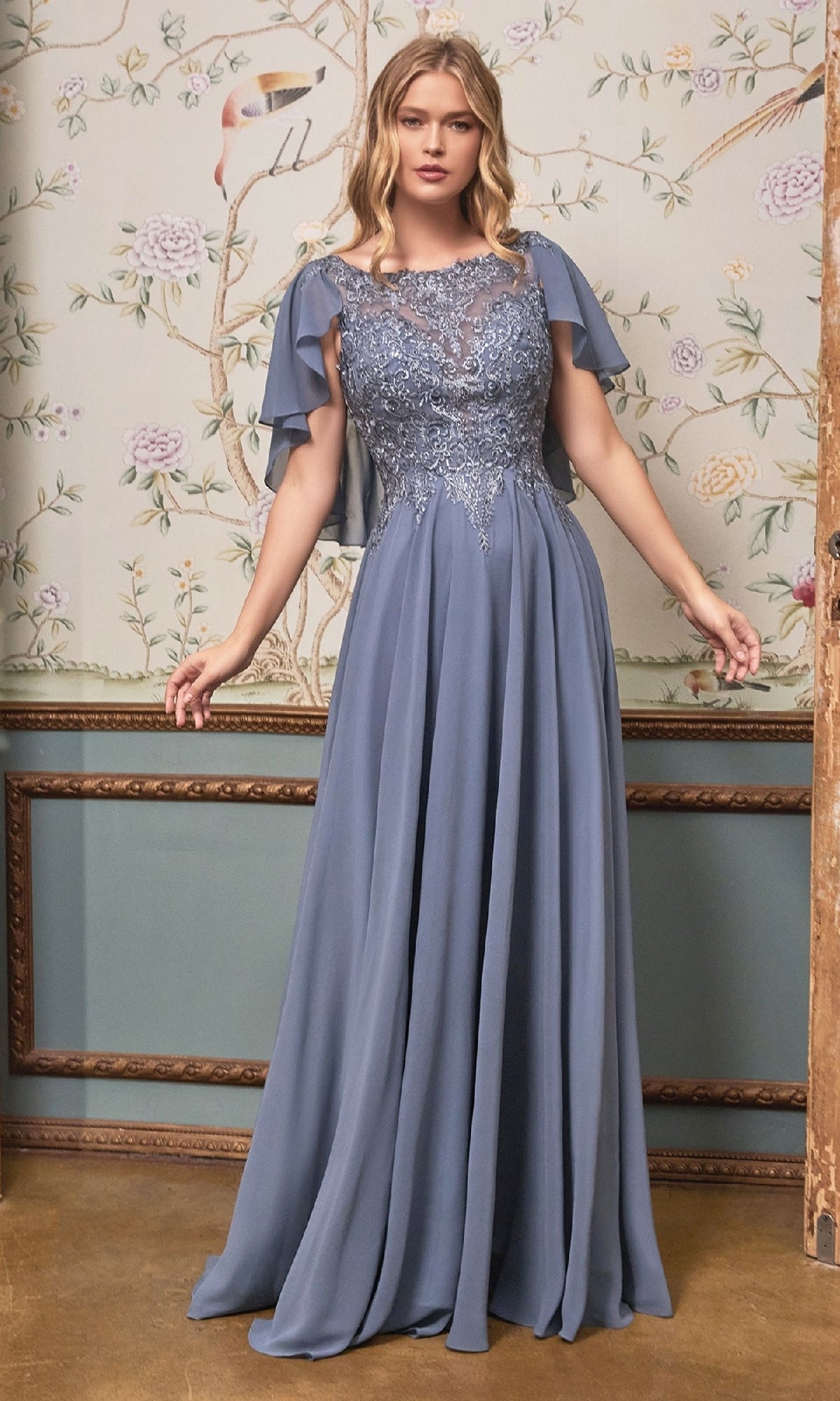A woman stands in front of floral wallpaper wearing the Cape-Sleeve Long A-Line Prom Dress: Ladivine HT101, a floor-length blue gown with lace bodice detailing and cape sleeves.